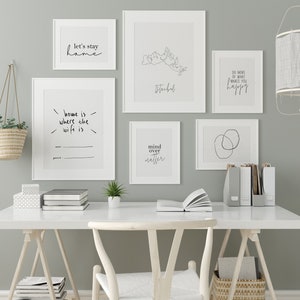 以下が含まれることがあります： ミニマリストデザインとインスピレーションを与える引用句が特徴の、額装されたアートプリントのコレクション。白いフレームに入ったプリントには、「let's stay home」や「mind over matter」などのフレーズが含まれています。アートワークは、白い机の上に展示されています。