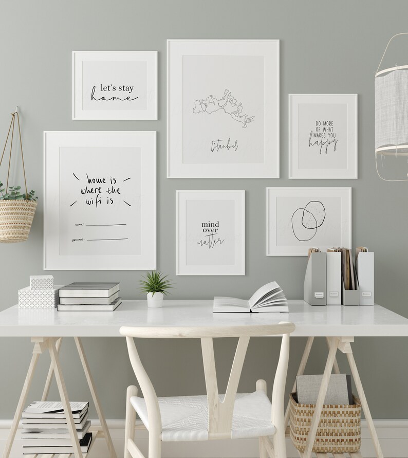 以下が含まれることがあります： ミニマリストデザインとインスピレーションを与える引用句が特徴の、額装されたアートプリントのコレクション。白いフレームに入ったプリントには、「let's stay home」や「mind over matter」などのフレーズが含まれています。アートワークは、白い机の上に展示されています。