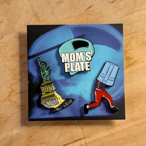 May include: Three enamel pins on a blue background. The pins are of a blue and white helmet with the text "MOM'S PLATE", a green Statue of Liberty with the text "GROUNDED FOR LIFE", and a red and blue figure with a blue shirt on its head.