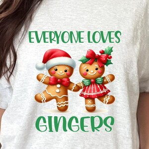May include: A light gray t-shirt featuring a graphic of two gingerbread people, one wearing a Santa hat and the other a red dress. The text "EVERYONE LOVES GINGERS" is printed in green. The shirt is worn by a person.