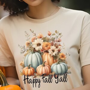 May include: A cream-coloured t-shirt with a watercolour design of pumpkins and flowers in shades of orange, peach, and blue. The text "Happy Fall Y'all" is below the design. The shirt is part of a seasonal collection.