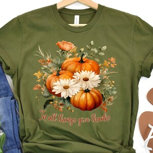 以下が含まれることがあります： かぼちゃとデイジーの花柄がプリントされた緑色のTシャツ。Tシャツには「In all things give thanks」という文字がプリントされています。