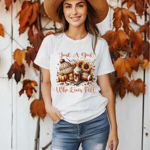 May include: White t-shirt featuring a graphic design of a cosy autumn scene. The design includes a pumpkin, a coffee cup, a book, a football, and a sunflower. The text on the shirt reads "Just a Girl Who Loves Fall".