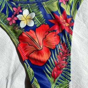 May include: A blue swimsuit bottom with a tropical floral print featuring red hibiscus flowers, green leaves, and white flowers. The fabric has a shiny, satin-like finish.