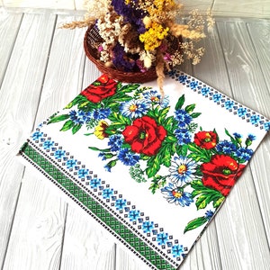 May include: White tablecloth with a floral pattern featuring red poppies, blue and white daisies, and green leaves. The border is a green and blue geometric pattern.