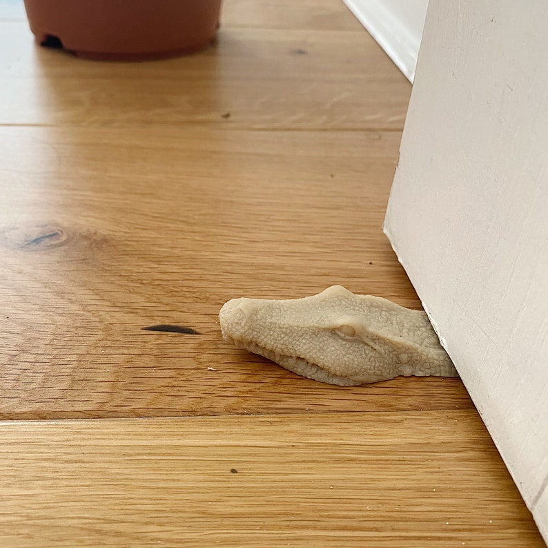 May include: A beige alligator-shaped doorstop with a textured surface. The doorstop is positioned under a white door.