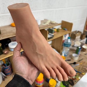 Könnte beinhalten: Ein realistisches, hautfarbenes Fußmodell aus Silikon. Der Fuß ist so positioniert, als würde er von einer Hand gehalten, mit leicht gekrümmten Zehen.