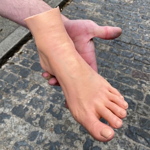 Puede incluir: Un pie protésico, de color carne, sostenido en una mano. El pie muestra detalles de venas y uñas de los pies. El fondo es una calle adoquinada.