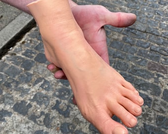 May include: A prosthetic foot, flesh-toned, held in a person's hand. The foot is detailed with visible veins and toenails. The background is a cobblestone street.