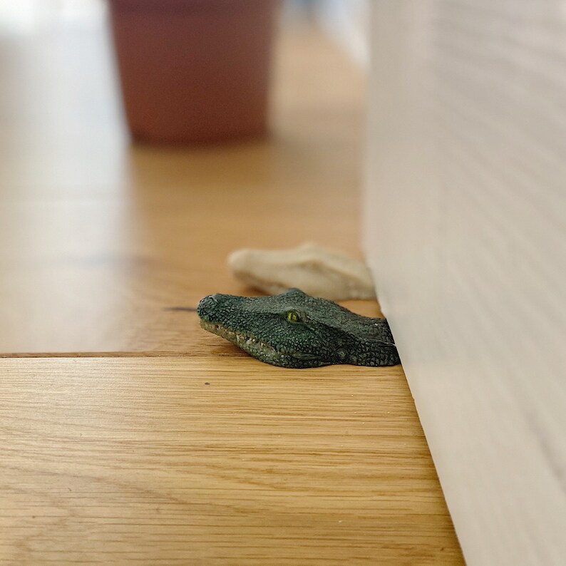 May include: A green alligator head doorstop with a yellow eye, positioned under a white door frame.