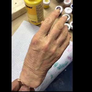 Op de afbeelding: Een close-up van een realistisch ogende hand gemaakt van siliconen. De hand is bleek en heeft zichtbare aderen en rimpels. De hand wordt vastgehouden door een hand van een persoon.