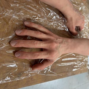 Pode incluir: Um close-up de duas mãos, uma com os dedos estendidos e a outra com os dedos fechados em punho. As mãos estão cobertas com um filme plástico transparente e estão apoiadas em uma superfície de madeira.