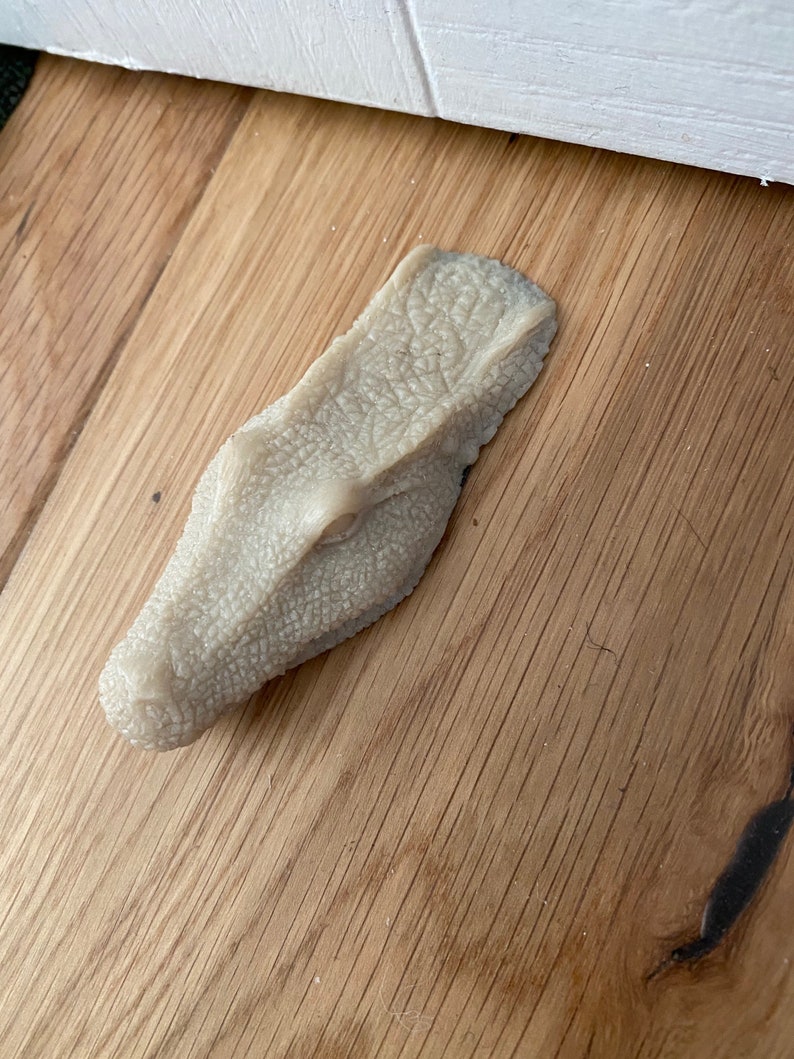 May include: A light brown, textured, alligator head doorstop. The doorstop is made of a soft, pliable material and is designed to prevent doors from slamming.