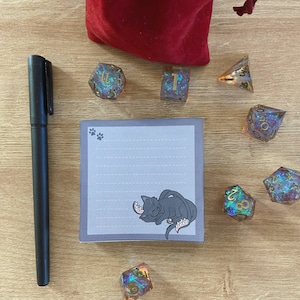 May include: A gray notepad with lined paper and a black cat illustration. The notepad is surrounded by iridescent dice with gold numbers. The dice are on a wooden surface.