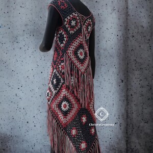 Peut inclure: Un gilet sans manches en crochet fait à la main, avec un motif géométrique gris, rouge et blanc. Le gilet est orné de franges en bas et d'un détail de dos lacé. Le gilet est présenté sur un mannequin.