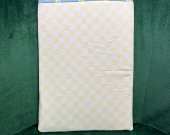 May include: A rectangular fabric item with a yellow and white checkered pattern. The top edge features a light blue fabric with small white daisy flowers. The item is resting on a dark green surface.