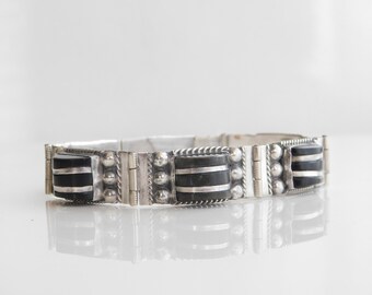 May include: A silver-toned bracelet with rectangular black and silver accents. The bracelet features a series of connected rectangular black stones with silver stripes, interspersed with silver beaded and textured elements. The bracelet is displayed on a white surface.
