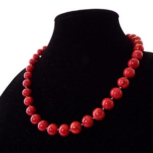 May include: A red beaded necklace displayed on a black velvet bust. The necklace features large, round beads in a vibrant red hue, strung together. The beads are evenly sized and closely spaced.