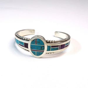 May include: A silver bracelet with a turquoise and purple inlay design. The bracelet has a round center piece with a geometric pattern.