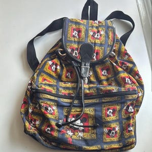May include: A vintage backpack featuring a repeating pattern of Mickey Mouse in gold frames on a navy blue background. The backpack has black straps, a drawstring closure, and a black leather flap with a buckle. The bag has a zippered front pocket.