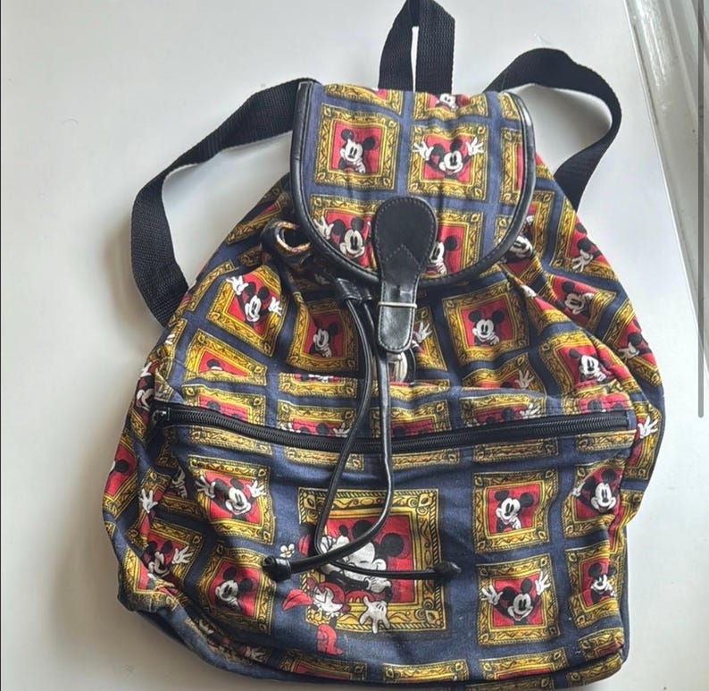 May include: A vintage backpack featuring a repeating pattern of Mickey Mouse in gold frames on a navy blue background. The backpack has black straps, a drawstring closure, and a black leather flap with a buckle. The bag has a zippered front pocket.