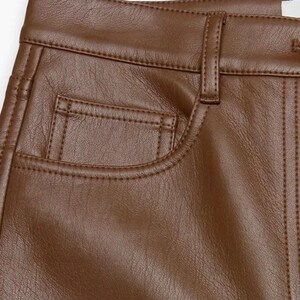 Può includere: Primo piano di pantaloni marroni in ecopelle. I pantaloni presentano un classico design a cinque tasche con cuciture a vista e chiusura con bottone. La consistenza del materiale è liscia e il colore è un marrone caldo e ricco.