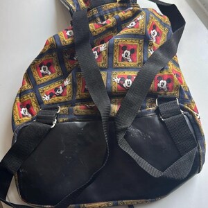 May include: A vintage backpack featuring a repeating pattern of Mickey Mouse in ornate frames. The backpack has black straps and a black leather-like bottom. The background is a dark blue with gold and red accents.