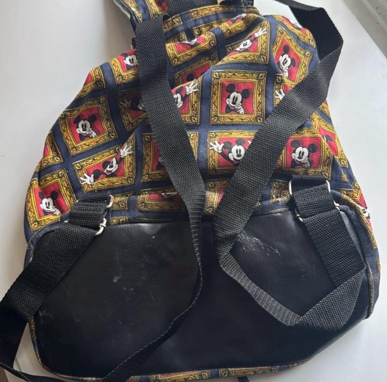 May include: A vintage backpack featuring a repeating pattern of Mickey Mouse in ornate frames. The backpack has black straps and a black leather-like bottom. The background is a dark blue with gold and red accents.