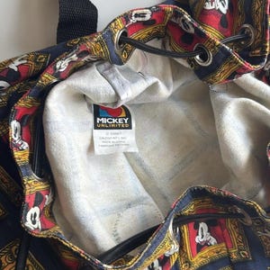 May include: A drawstring backpack with a repeating pattern of Mickey Mouse in ornate frames on a navy blue background. The interior is a light denim color. The tag reads "Mickey Unlimited".