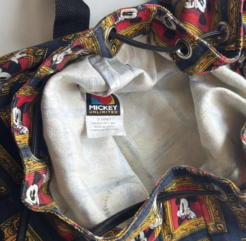 May include: A drawstring backpack with a repeating pattern of Mickey Mouse in ornate frames on a navy blue background. The interior is a light denim color. The tag reads "Mickey Unlimited".