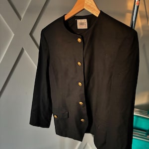 Op de afbeelding: Een zwarte blazer met gouden knopen en een ronde hals hangt aan een houten hanger. De blazer heeft een klassiek ontwerp met een getailleerde pasvorm, met een kleine zak aan elke kant. Het label luidt "CORRO C."