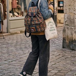 May include: A patterned backpack with a drawstring closure and a buckle, paired with a canvas tote bag. The tote bag has the text "REUSABLE COTTON BOOKSTORE". The person is wearing a denim jacket, corduroy pants, and black high-top sneakers.