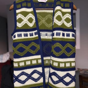 May include: A vintage open-front vest with a geometric pattern. The vest has navy blue trim and accents, with olive green and cream-coloured geometric shapes. The vest is hanging on a wooden hanger.