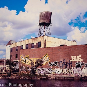 以下が含まれることがあります： 壁に落書きが描かれたレンガ造りの建物。落書きには、黄色の犬、"Art Cash!"と"Bug."という文字が描かれている。建物の頂上には、給水塔がある。