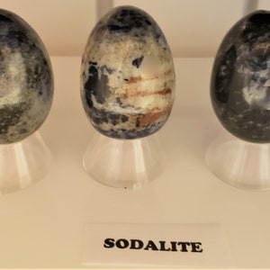 May include: Three smooth, polished sodalite eggs, each with a unique pattern of blue, white, and gray, are displayed on individual stands. The label below reads "SODALITE".
