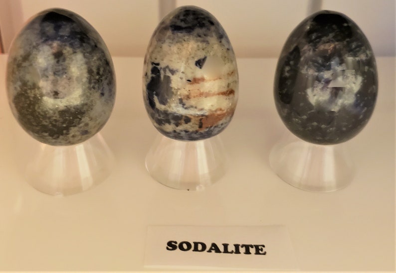 May include: Three smooth, polished sodalite eggs, each with a unique pattern of blue, white, and gray, are displayed on individual stands. The label below reads "SODALITE".