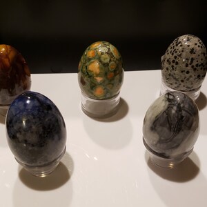 May include: Five polished stone eggs, each with a unique pattern and color, displayed on individual stands. The eggs are a mix of brown, green, blue, gray, and black.