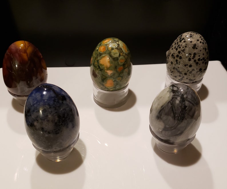 May include: Five polished stone eggs, each with a unique pattern and color, displayed on individual stands. The eggs are a mix of brown, green, blue, gray, and black.