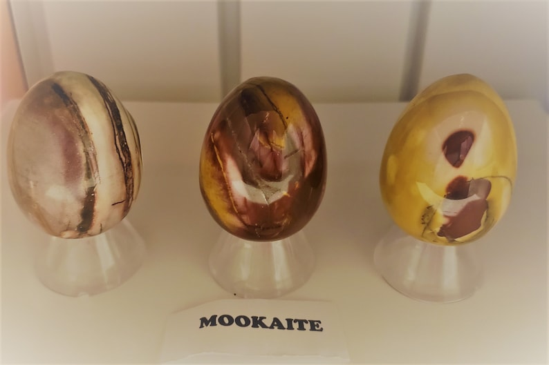 May include: Three polished Mookaite eggs, each with a unique pattern of brown, yellow, and white stripes and swirls, displayed on clear stands. The label below reads "MOOKAITE".