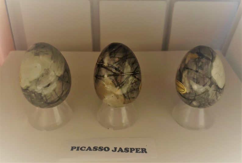 May include: Three smooth, egg-shaped stones with a gray and white marbled pattern. The stones are displayed on small white stands. The text "PICASSO JASPER" is printed below the stones.