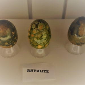 May include: Three green, brown, and white rhyolite stones on white stands. The stones are polished and have a speckled pattern. The label below the stones reads "Rhyolite".