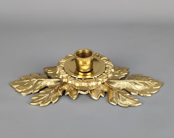 May include: Gold metal candle holder with a floral design and a round base. The holder has five leaves that extend out from the center. The center of the holder has a round base with a small cup for the candle.