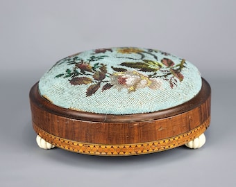 May include: A round beaded footstool with a floral design on the top. The footstool has a wooden base with a marquetry inlay and four white wooden feet.