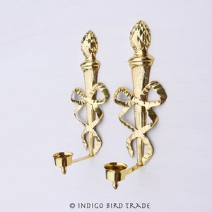 May include: A pair of gold metal wall sconces with a torch design and a ribbon detail. Each sconce has a candle holder.