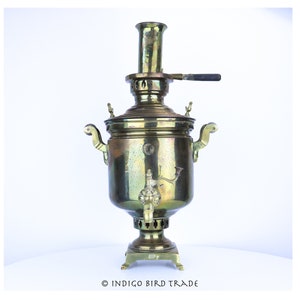 May include: A vintage brass samovar with a traditional design. The samovar has a spout and a handle on the side, and a tall chimney on top. The samovar is sitting on a white surface.