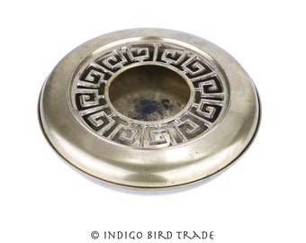 May include: A round brass incense burner with a Greek key design. The burner has a raised rim and a central opening for incense.