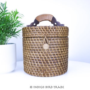 May include: A brown woven wicker basket with a wooden handle and a brown leather closure. The basket is round and has a lid.