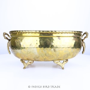 May include: A gold oval planter with hammered texture and ornate handles. The planter has three decorative feet.