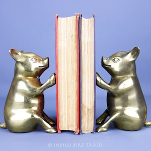 Könnte beinhalten: Ein Paar Messing-Buchstützen in Form von Schweinen, die ein Buch mit rotem und violettem Rücken halten. Die Buchstützen sitzen mit eingerollten Schwänzen.