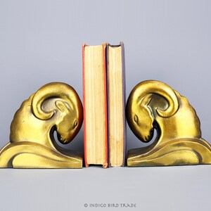 Könnte beinhalten: Ein Paar goldfarbene Buchstützen in Form von Widdern mit gesenktem Kopf. Die Buchstützen halten ein Buch mit rotem Rücken und gelbem Einband.